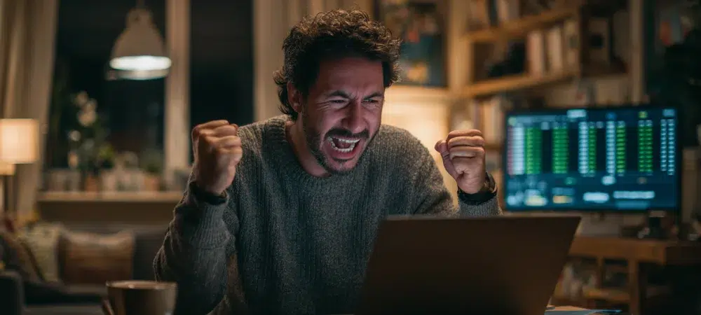 Un hombre sentado en un escritorio en una casa acogedora, celebrando después de ganar una gran apuesta deportiva en línea en su computadora portátil